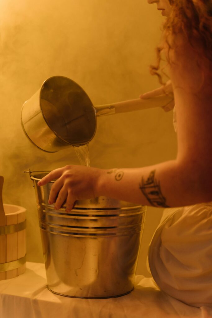 pexels photo 8284386 8284386 A woman with a tattoo pours water into a stainless steel bucket in a steamy setting.